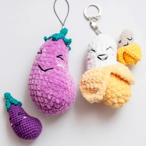 May include: Three crocheted plush keychains shaped like a banana, eggplant, and a smaller eggplant. The banana keychain is white with a yellow peel and has a happy face. The larger eggplant keychain is purple with a green stem and a happy face. The smaller eggplant keychain is purple with a green stem and a happy face.
