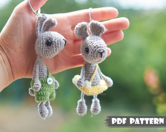 May include: Two crocheted bunny keychains, one wearing green overalls and the other a yellow skirt. Both bunnies have gray fur and black eyes. The keychains are held in a hand against a green background. PDF PATTERN.
