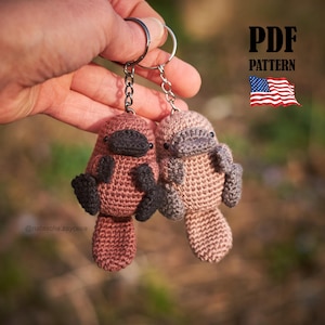 May include: Two crocheted platypus keyrings in shades of brown and grey, each with a black beak and feet. The keyrings are attached to silver key rings. The image includes the text "PDF PATTERN" and a waving American flag.