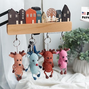 May include: Five crocheted reindeer keychains hanging from hooks on a wooden shelf. The reindeer are in various colours, including pink, blue, and brown. The shelf is decorated with miniature houses and a small tree.