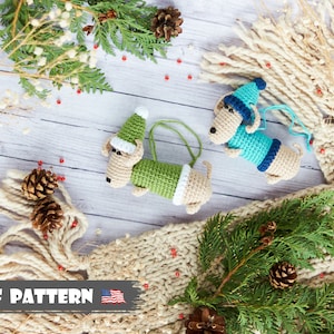 Op de afbeelding: Twee gehaakte teckel-versieringen, een in groen en een in blauw, met kerstmanmutsen. De versieringen liggen op een witte houten achtergrond met dennentakken en een witte gebreide sjaal.