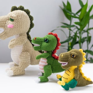 May include: Three crocheted dinosaur toys. The largest dinosaur is beige with green accents. The middle dinosaur is green with red accents. The smallest dinosaur is green with yellow accents.