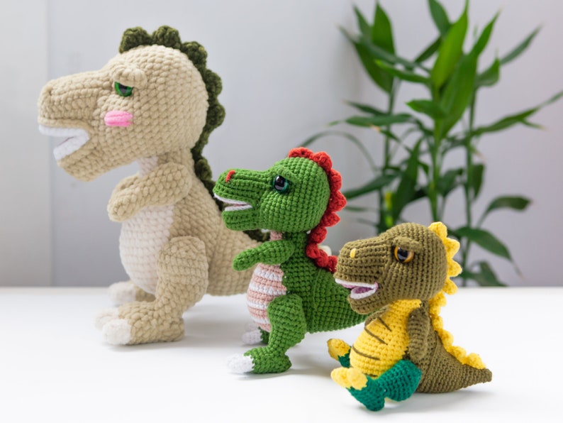May include: Three crocheted dinosaur toys. The largest dinosaur is beige with green accents. The middle dinosaur is green with red accents. The smallest dinosaur is green with yellow accents.