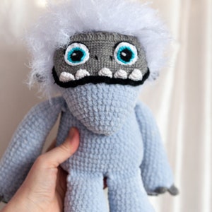 May include: A crocheted plush toy resembling a yeti with a white fur head, blue body, and gray claws. The toy has large blue eyes and a black mouth with white teeth.