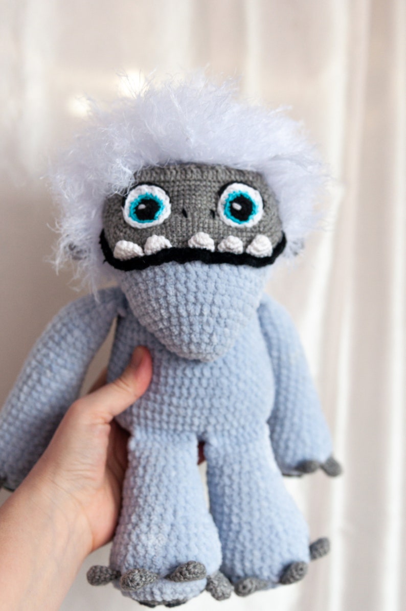 May include: A crocheted plush toy resembling a yeti with a white fur head, blue body, and gray claws. The toy has large blue eyes and a black mouth with white teeth.