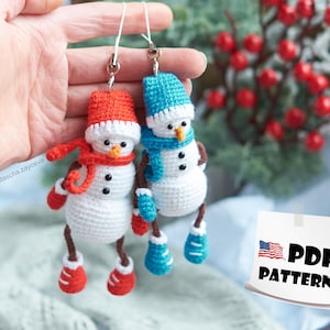 Puede incluir: Dos adornos de muñeco de nieve de crochet con sombreros, bufandas y guantes rojos y azules. Los adornos están colgados de una cuerda y se sostienen en una mano. El texto "PDF PATTERN" está en un papel blanco con una pequeña bandera estadounidense.