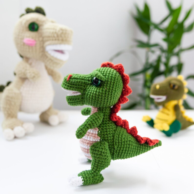 May include: Three crocheted dinosaur toys. One is green with red spikes and white feet, another is beige with pink cheeks, and the third is green and yellow. The dinosaurs are on a white surface.