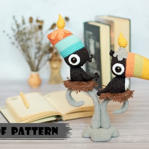 Puede incluir: Dos tucanes de crochet con picos y sombreros coloridos posados en una rama de árbol de crochet. Los tucanes están sentados sobre una superficie de madera con un libro y un lápiz en el fondo.