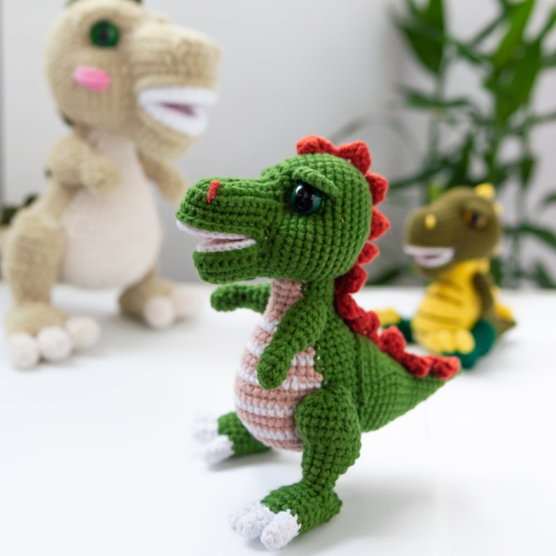 May include: Three crocheted dinosaur toys. One is green with red accents and pink stripes, one is beige with pink accents, and one is yellow with green accents.