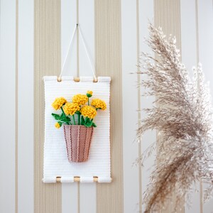 May include: A white crocheted wall hanging with a yellow crocheted flower bouquet in a pink crocheted pot.