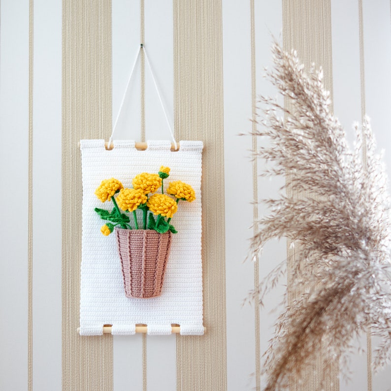 May include: A white crocheted wall hanging with a yellow crocheted flower bouquet in a pink crocheted pot.