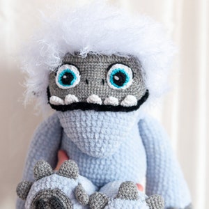 May include: A crocheted plush toy resembling a yeti with a white fluffy head, blue body, and gray feet. The toy has large blue eyes and a black mouth.
