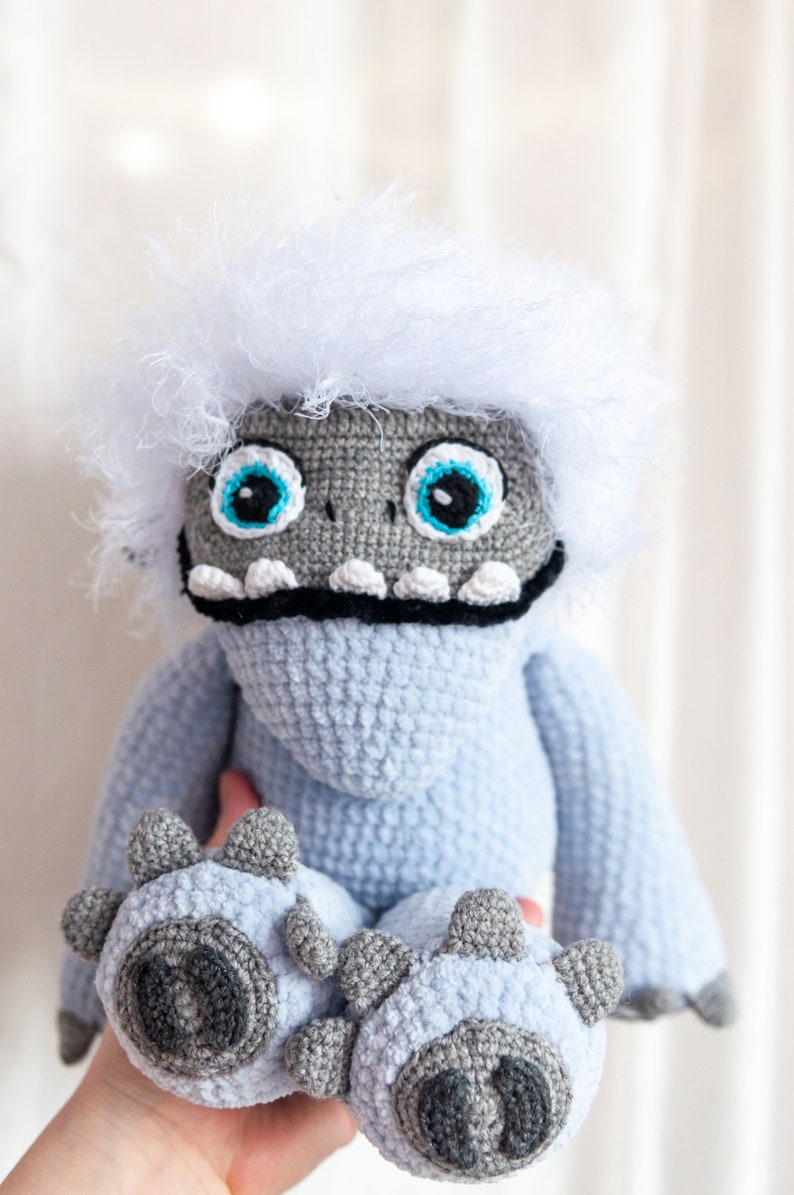 May include: A crocheted plush toy resembling a yeti with a white fluffy head, blue body, and gray feet. The toy has large blue eyes and a black mouth.