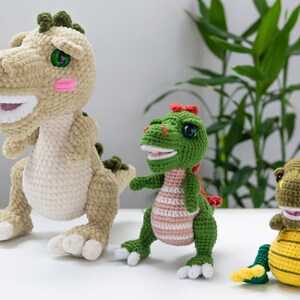 May include: Three crocheted dinosaur toys. The largest dinosaur is beige with green accents and has a toothy grin. The middle dinosaur is green with pink stripes and a red crest. The smallest dinosaur is yellow with green accents and a toothy grin.