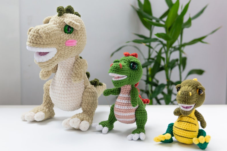 May include: Three crocheted dinosaur toys. The largest dinosaur is beige with green accents and has a toothy grin. The middle dinosaur is green with pink stripes and a red crest. The smallest dinosaur is yellow with green accents and a toothy grin.