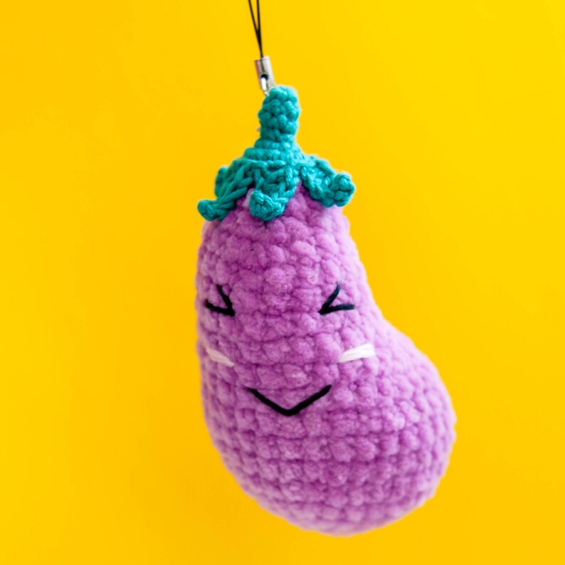 May include: A purple crocheted eggplant with a green crocheted stem and a smiling face. The eggplant is hanging from a string.