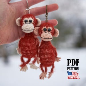 May include: Two handmade crochet monkey keychains. The monkeys are red with beige faces, ears, and hands. They have black eyes and are attached to metal keyrings. The image also includes the text "PDF PATTERN" and an American flag.