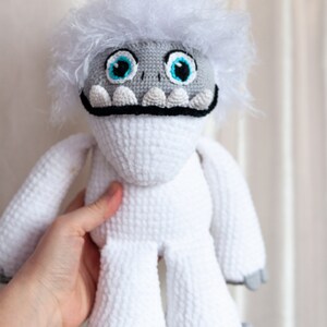 May include: White crocheted yeti plush toy with blue eyes and a black mouth. The toy has a fluffy white fur and gray feet.