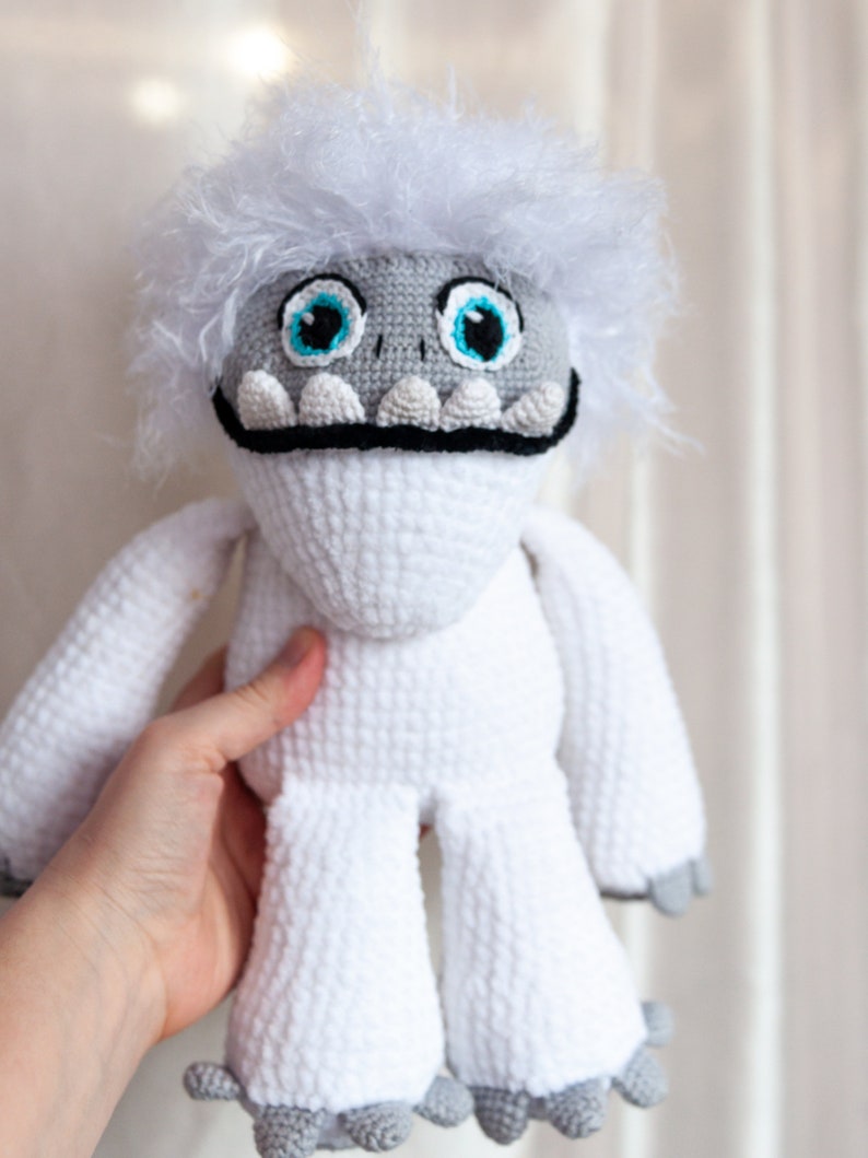 May include: White crocheted yeti plush toy with blue eyes and a black mouth. The toy has a fluffy white fur and gray feet.