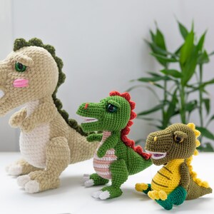 May include: Three crocheted dinosaur toys. The largest dinosaur is beige with green accents. The middle dinosaur is green with red accents. The smallest dinosaur is green and yellow with a light green belly.