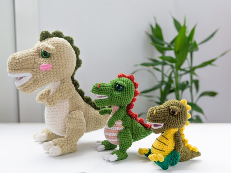 May include: Three crocheted dinosaur toys. The largest dinosaur is beige with green accents. The middle dinosaur is green with red accents. The smallest dinosaur is green and yellow with a light green belly.
