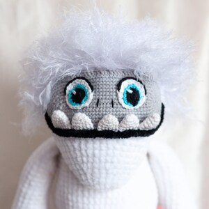 May include: A white crocheted plush toy with a gray face, blue eyes, and a black mouth. The toy has a white fluffy hair.