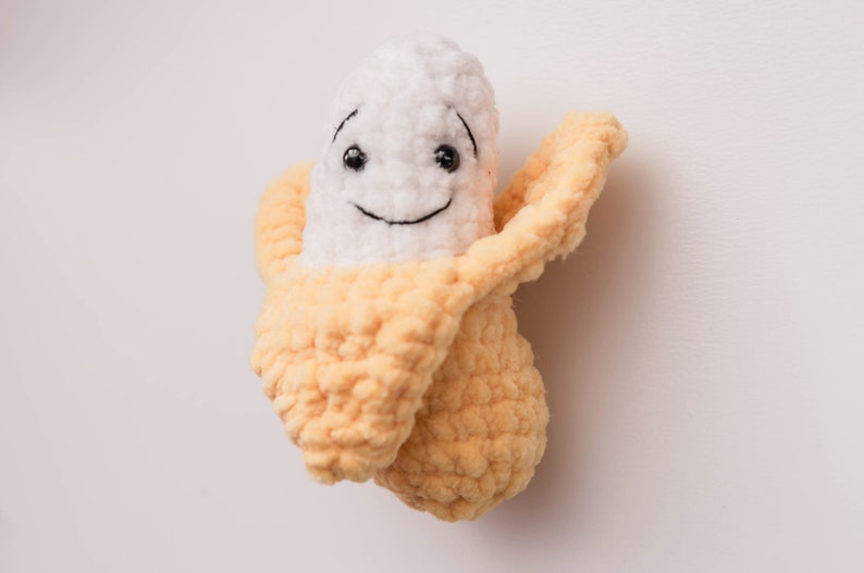 May include: A crocheted banana plush toy with a smiling face. The banana is white with a yellow peel.