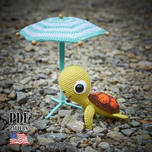 Pode incluir: Um brinquedo de tartaruga de crochê com corpo verde-claro e casco marrom e laranja, posicionado sob um pequeno guarda-sol de praia de crochê com listras azul-claras e brancas e detalhes em contas de madeira. A imagem inclui o texto PDF PATTERN.