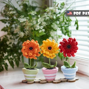 Op de afbeelding: Drie gehaakte bloemstukken in potten. De bloemen zijn oranje, geel en rood. De potten zijn groen, roze en blauw met witte strepen. De bloemen staan op een houten oppervlak.