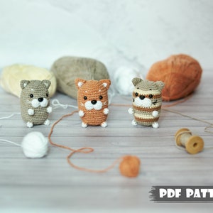 May include: Three crocheted animal plushies in brown, orange, and white. The plushies are sitting on a wooden surface with yarn balls and spools of yarn. The image includes the text "PDF PATTERN".