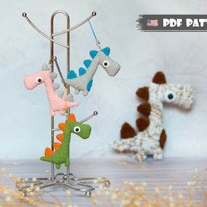 Può includere: Tre giocattoli di dinosauro all'uncinetto in rosa, verde e grigio appesi a un supporto in metallo. I dinosauri hanno occhi grandi e sono realizzati con una varietà di punti.  MODELLO PDF.