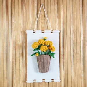May include: A white crocheted wall hanging with a brown crocheted flower pot and yellow crocheted flowers.