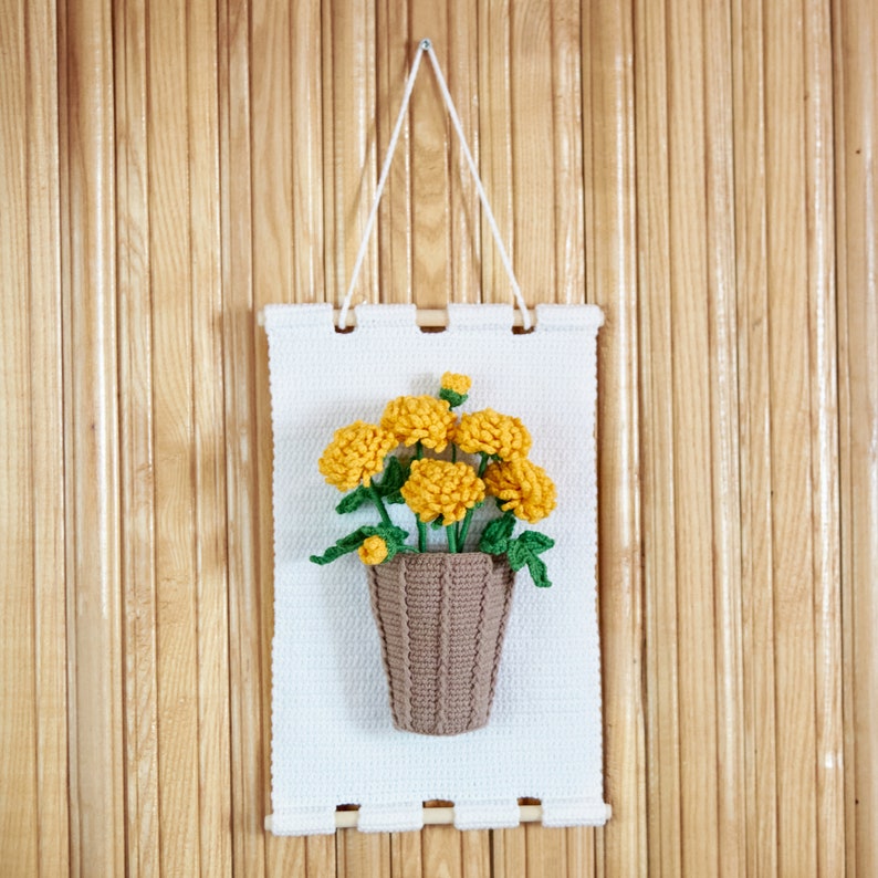 May include: A white crocheted wall hanging with a brown crocheted flower pot and yellow crocheted flowers.