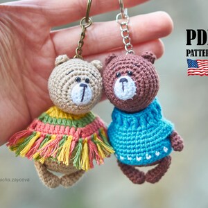 Puede incluir: Dos llaveros de oso de ganchillo hechos a mano. Un oso es de color beige con un poncho con flecos de colores, el otro es marrón con un suéter azul. Ambos tienen ojos negros y caras blancas. Los llaveros están unidos a anillos de metal. La imagen incluye el texto "PDF PATTERN" y una bandera estadounidense.