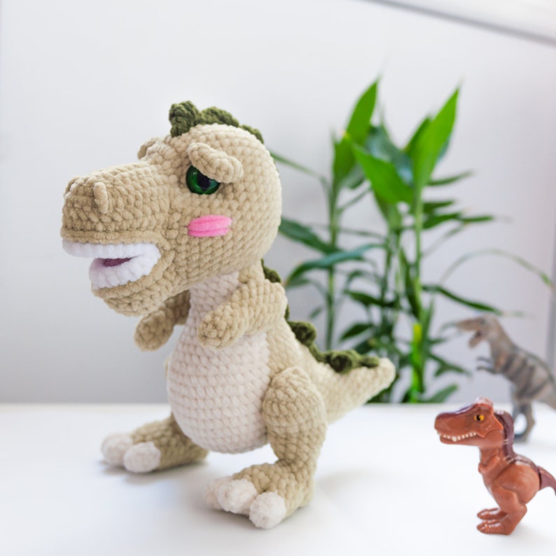 May include: A crocheted green and beige dinosaur toy with a pink blush on its cheeks. The dinosaur has a wide open mouth with white teeth and green eyes.