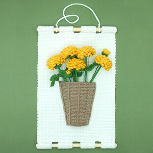 May include: A white crocheted wall hanging with a brown crocheted pot and yellow crocheted flowers.