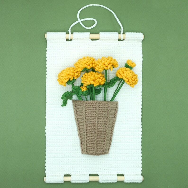 May include: A white crocheted wall hanging with a brown crocheted pot and yellow crocheted flowers.