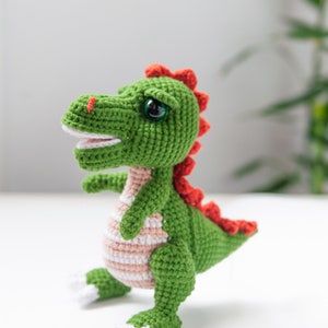 May include: A green crocheted dinosaur toy with red and white accents. The dinosaur has a wide smile and large eyes.