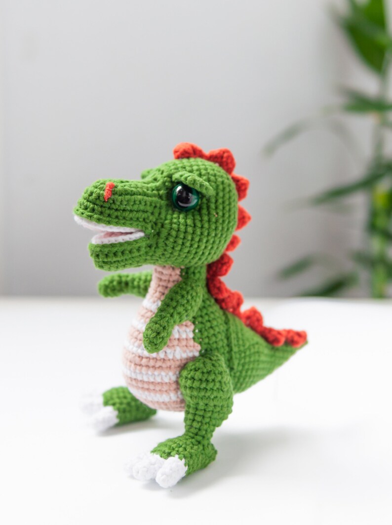 May include: A green crocheted dinosaur toy with red and white accents. The dinosaur has a wide smile and large eyes.