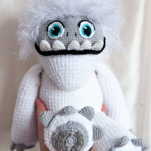 May include: White crocheted plush toy with a furry white head, blue eyes, and a black mouth. The toy has gray feet and is being held by a hand.