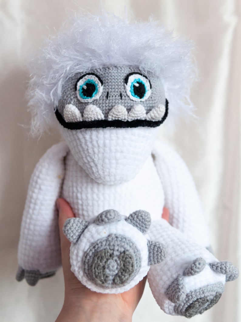 May include: White crocheted plush toy with a furry white head, blue eyes, and a black mouth. The toy has gray feet and is being held by a hand.