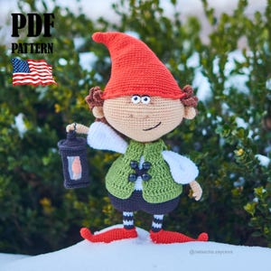 以下が含まれることがあります： 赤い帽子、緑のベスト、黒いショートパンツを着て、ランタンを持っている手作りのかぎ針編みのノーム人形。人形は茶色の髪で、赤と白のストライプの靴下と靴を履いています。画像には「PDF PATTERN」というテキストとアメリカ国旗が含まれています。