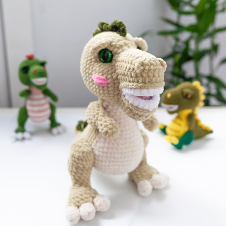 May include: A crocheted plush toy dinosaur with green eyes, pink cheeks, and white teeth. The dinosaur is standing on a white surface with two other smaller crocheted dinosaurs.
