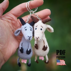 May include: Two crocheted goat keychains, one light gray and one cream, each featuring dark brown horns, small black hooves, and pink udders. The goats are attached to a silver metal keyring. The image includes the text PDF PATTERN and a US flag.