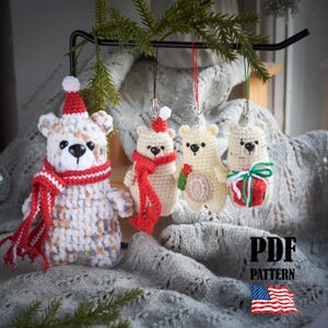 以下が含まれることがあります： お祝いの衣装を着た4つの編み物ベアオーナメント。大きなクマは赤と白のマフラーと帽子をかぶっています。小さなクマは赤いスカーフ、帽子、プレゼントの包装紙を持っています。オーナメントは黒い金属棒から吊り下げられています。