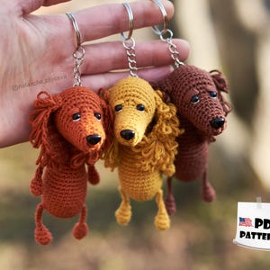 Może przedstawiać: Trzy breloczki z dzianiny w kształcie szczeniaka rasy dachshund w kolorze brązowym, żółtym i pomarańczowym. Breloczki wiszą na srebrnych kółkach. Obraz zawiera tekst "PDF PATTERN".