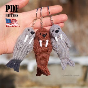 Op de afbeelding: Drie gehaakte walrus sleutelhangers in grijs en bruin, elk met witte slagtanden en zwarte ogen. De sleutelhangers zijn bevestigd aan zilveren ringen. De afbeelding bevat de tekst "PDF PATTERN" en een kleine Amerikaanse vlag.