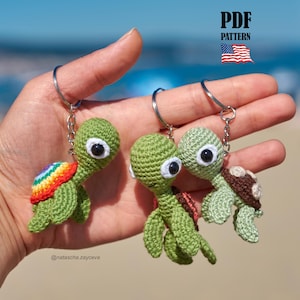 May include: Three small crocheted turtle keychains with large black and white eyes. One turtle has a rainbow-colored shell, one has a dark red shell, and one has a brown shell. The text PDF PATTERN and a small American flag icon are visible.