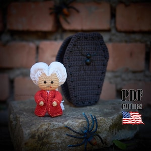 Puede incluir: Una muñeca de ganchillo con cabello blanco y un abrigo rojo está junto a un ataúd de ganchillo gris oscuro. La muñeca tiene una cara bronceada y lleva un abrigo rojo. Una araña azul y negra está en primer plano. Logotipo de patrón PDF.