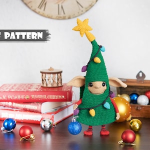 Puede incluir: Patrón de crochet para un juguete amigurumi de elfo de árbol de Navidad. El elfo lleva un disfraz de árbol de Navidad verde con una estrella amarilla en la parte superior y luces de colores. El elfo tiene una cara marrón y grandes ojos marrones.